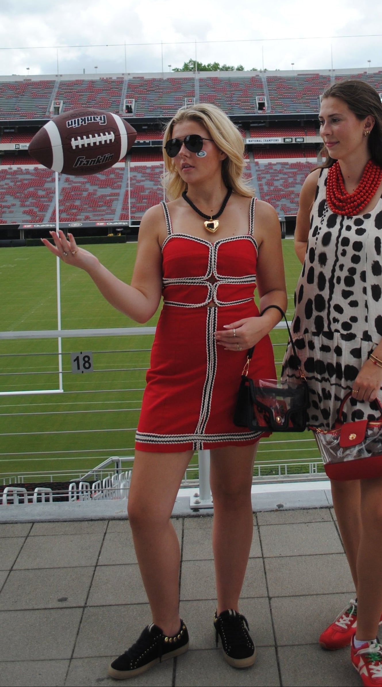 Two women standing in a stadium, one wearing a red mini dress with white trim and holding a football, and the other wearing a black-and-white spotted dress. A football is suspended in the air in front of them.