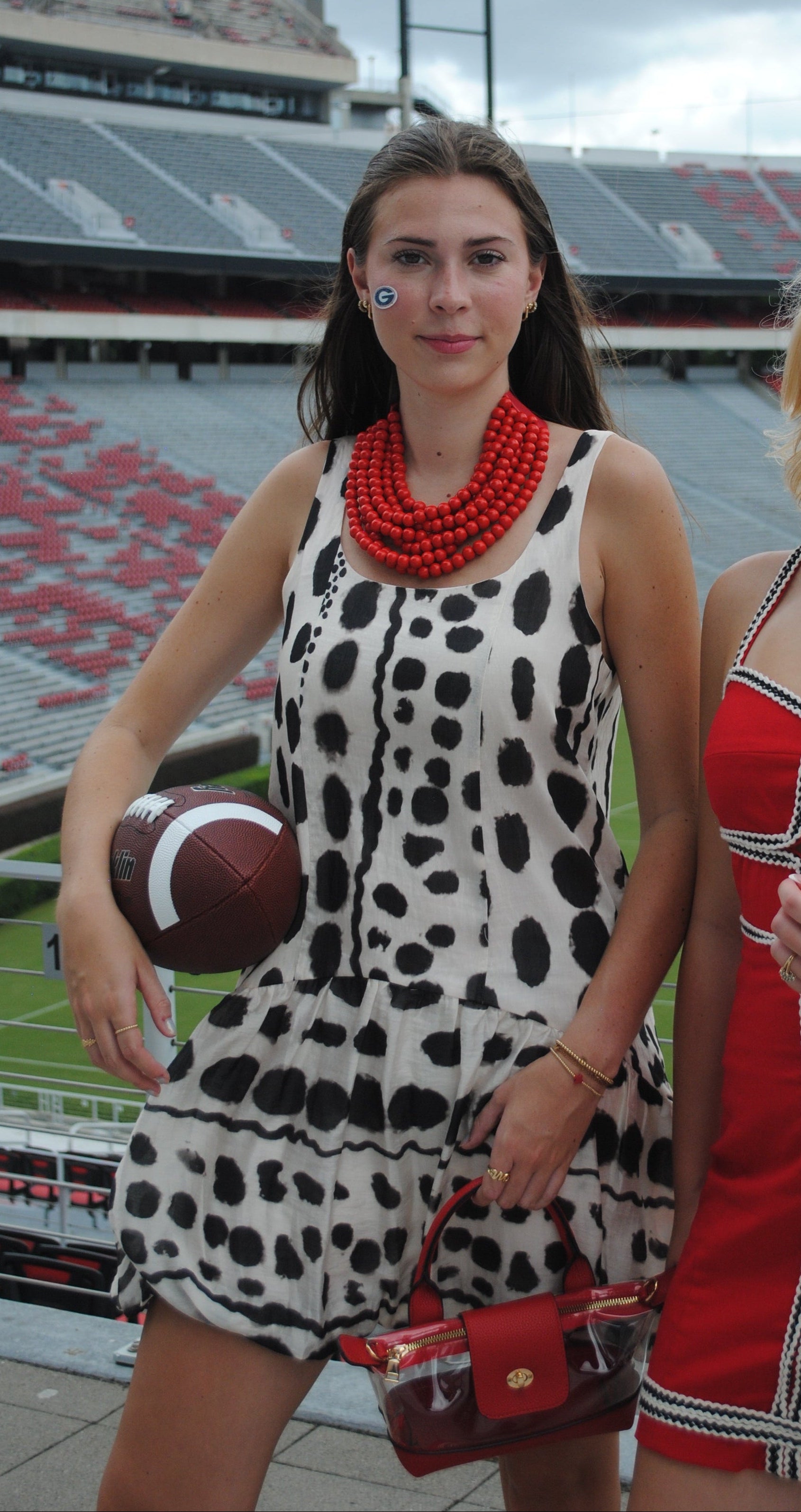 Two women posing in a stadium, one wearing a black and white cow-print dress with a red beaded necklace and holding a football, and the other wearing a red dress with black and white trim.