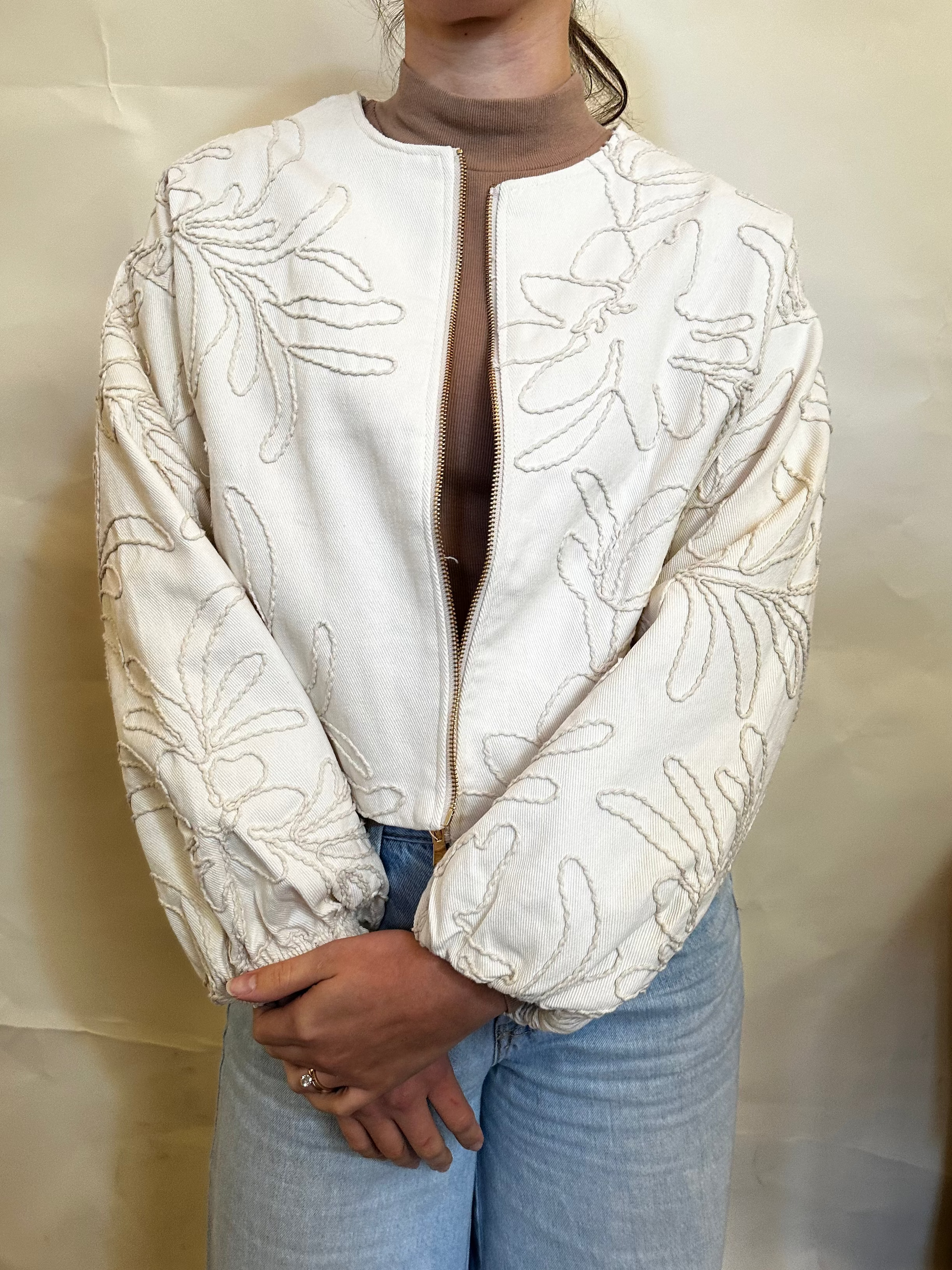 Person wearing a white jacket with leaf patterns and light blue jeans against a plain background