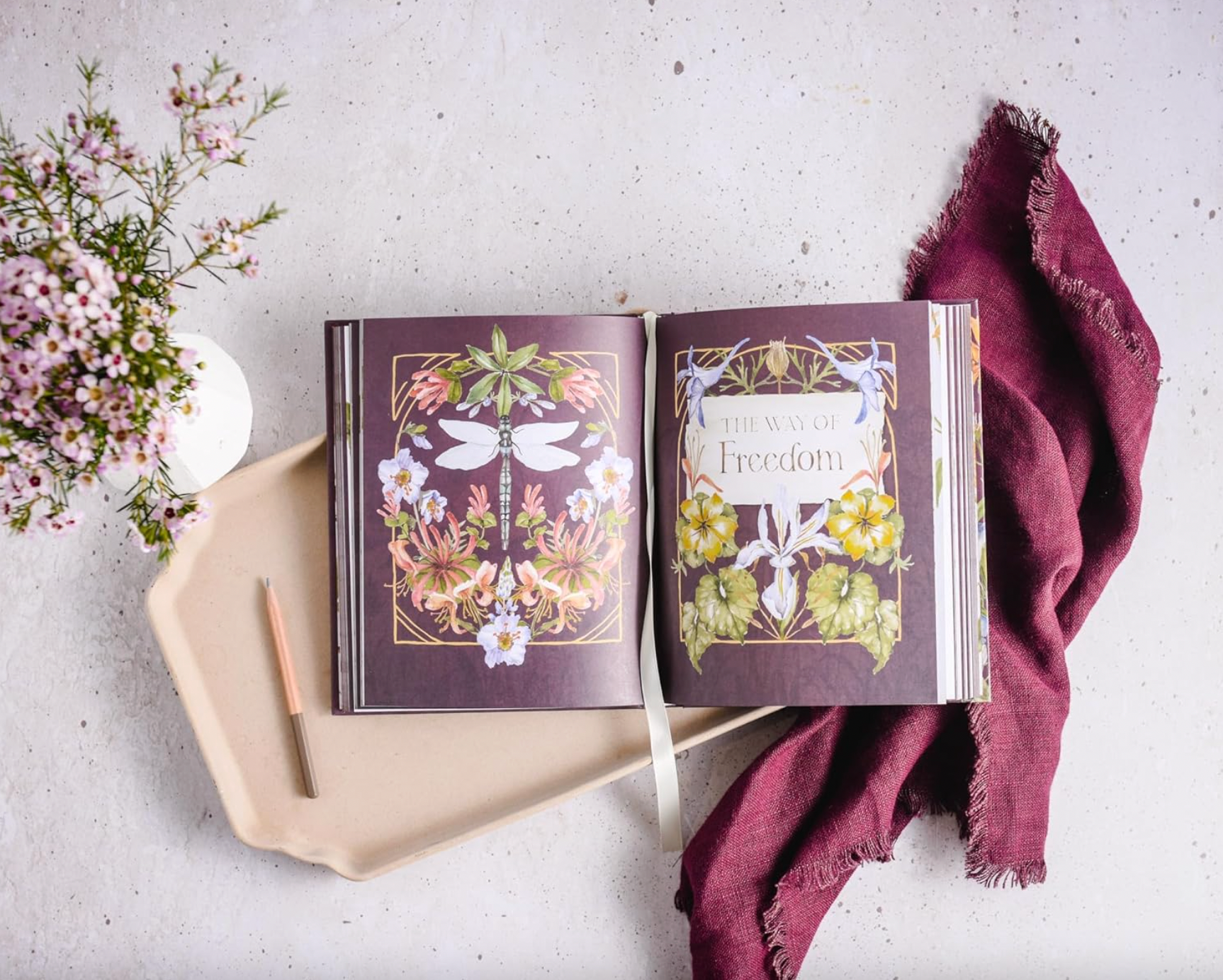Open book with floral designs on a light surface with flowers and a towel.