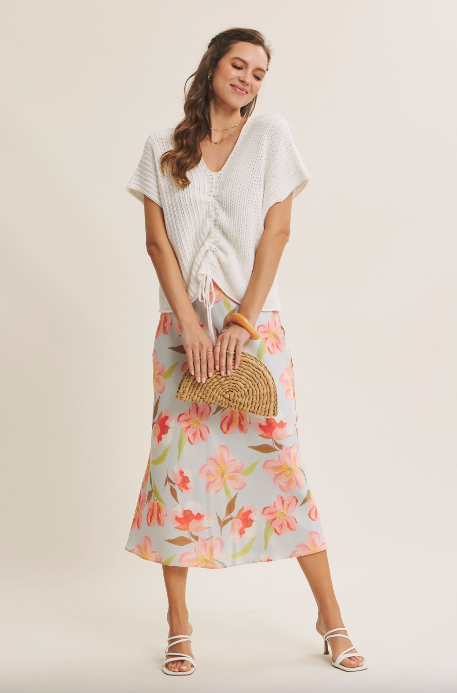 Woman wearing a white top and floral skirt with a woven clutch against a plain background