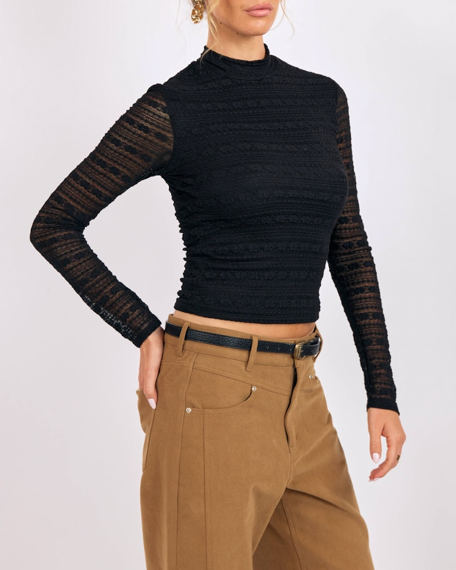 Person wearing a black ribbed long-sleeve top with sheer sleeves and beige pants on a white background