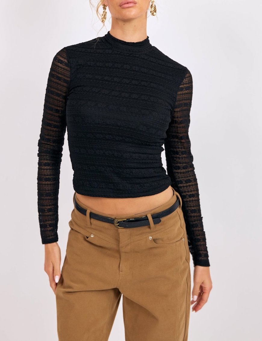 Person wearing a black ribbed long-sleeve top with sheer sleeves and beige pants on a white background