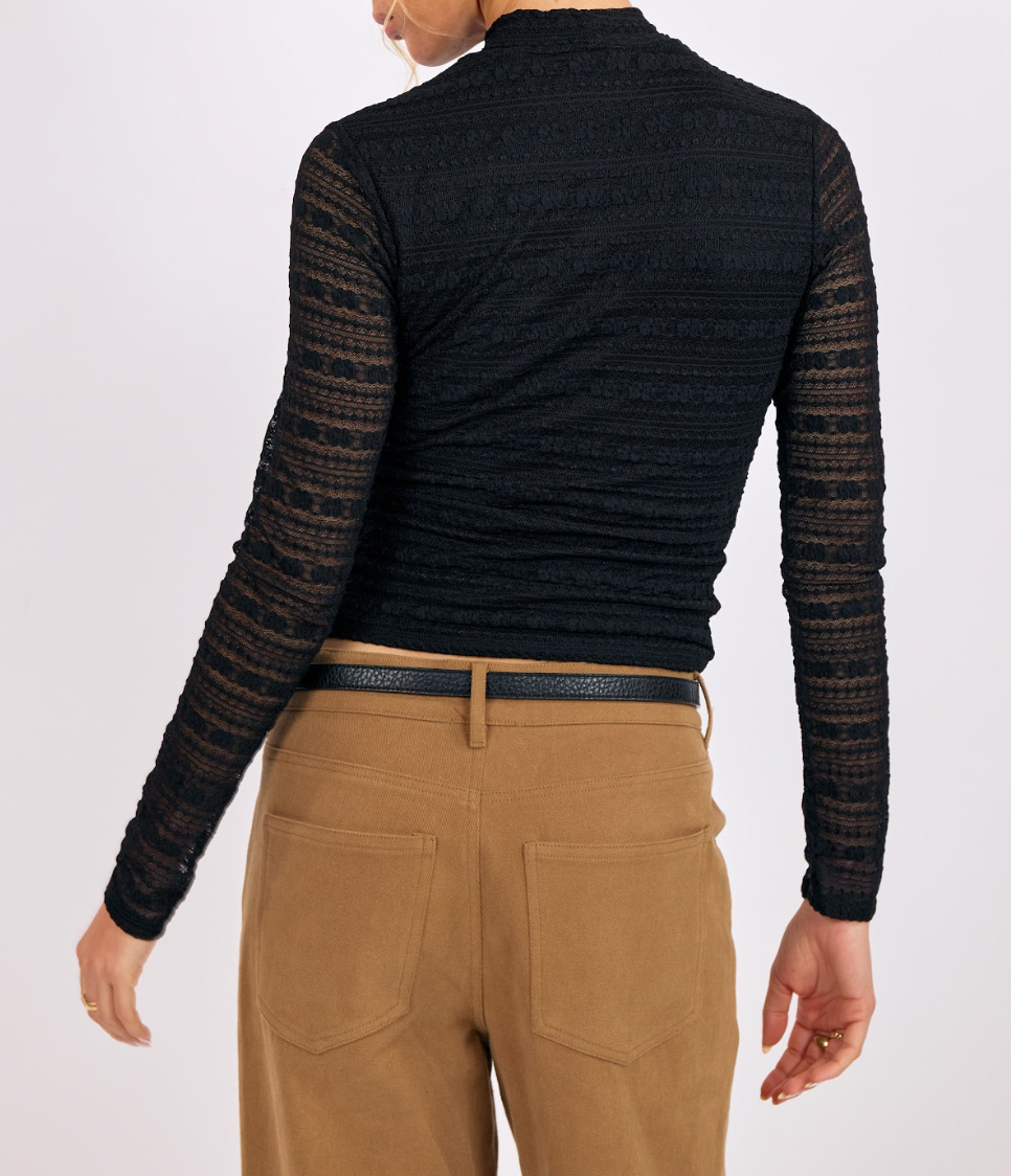 Person wearing a black long-sleeve top with sheer sleeves and beige pants on a white background