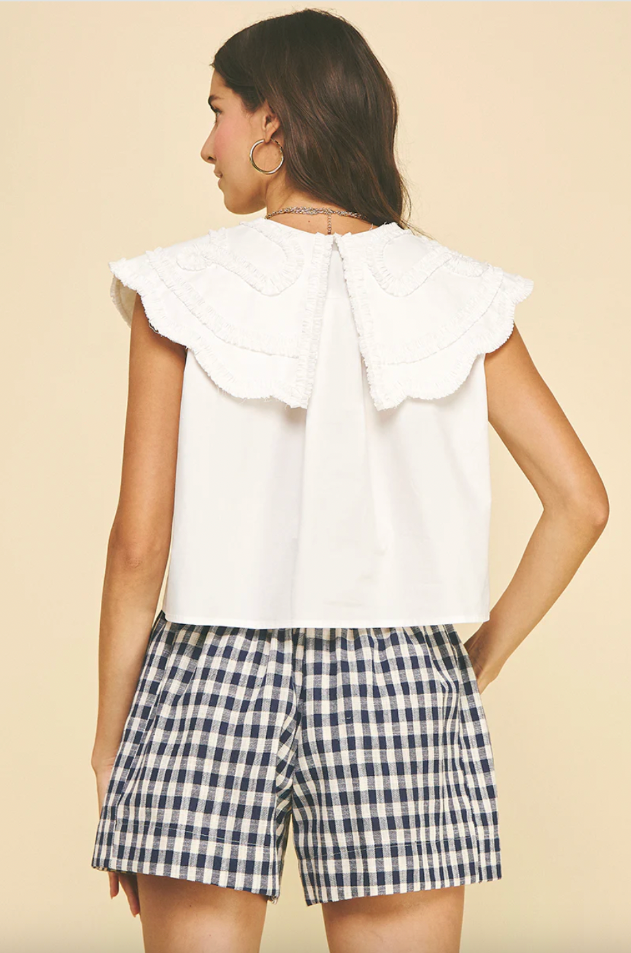 Woman wearing a white top with ruffled collar and plaid shorts on a beige background