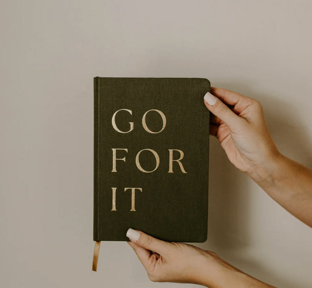 Hand holding a dark green notebook with 'GO FOR IT' text on a plain background