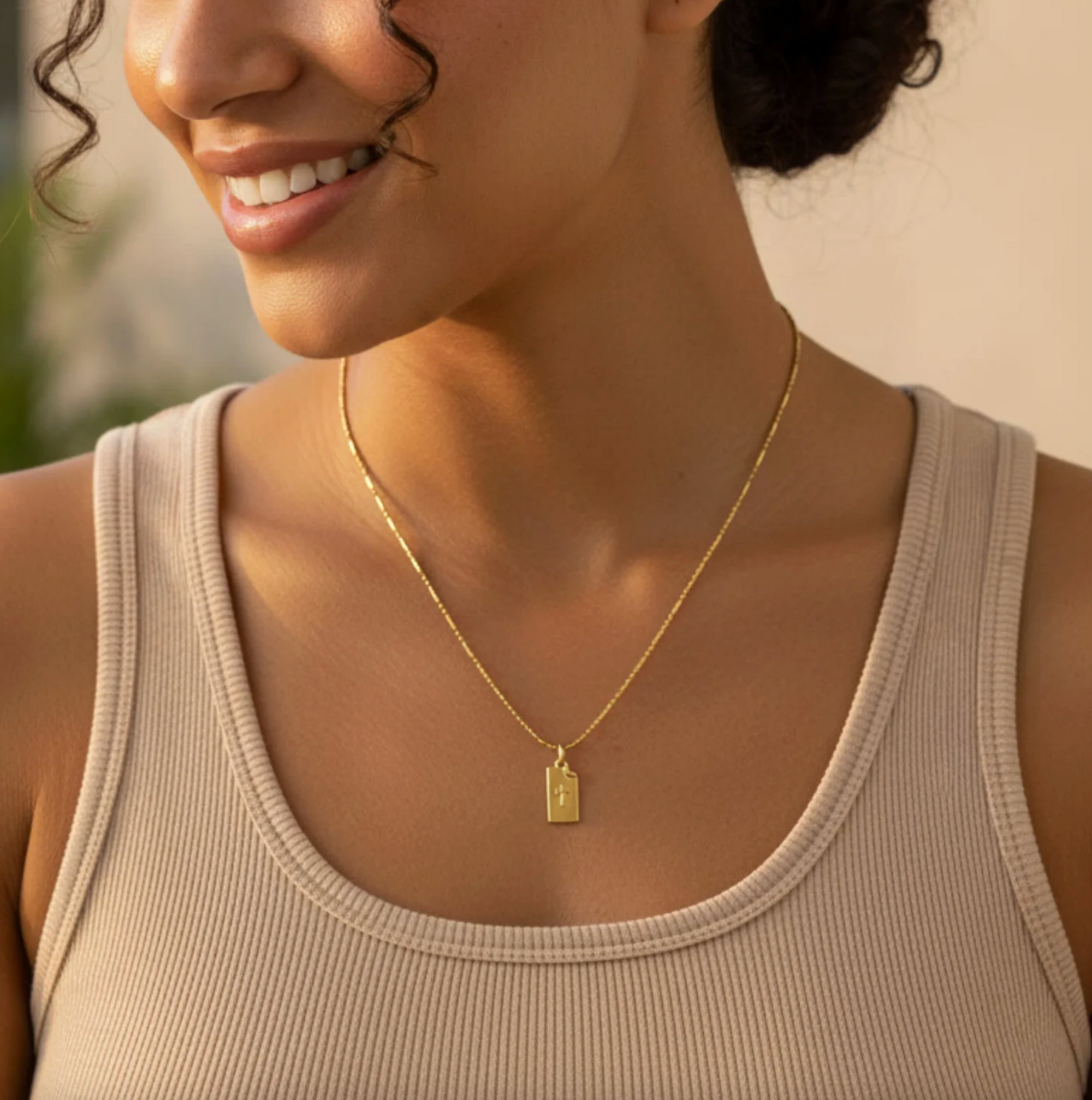 Woman wearing a gold necklace with a tag pendant, smiling outdoors.