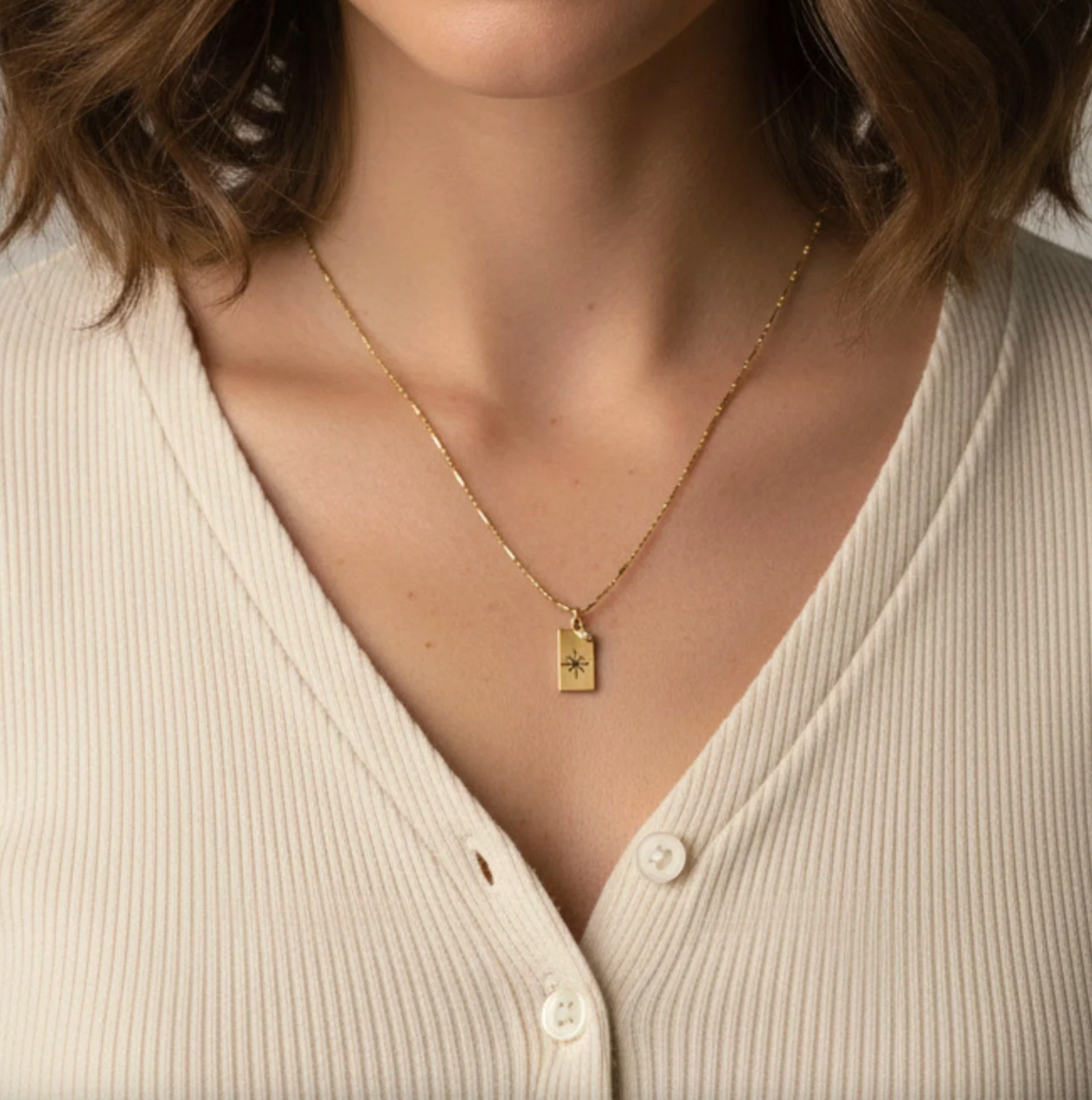 Gold necklace with a rectangular pendant worn by a person in a beige top.