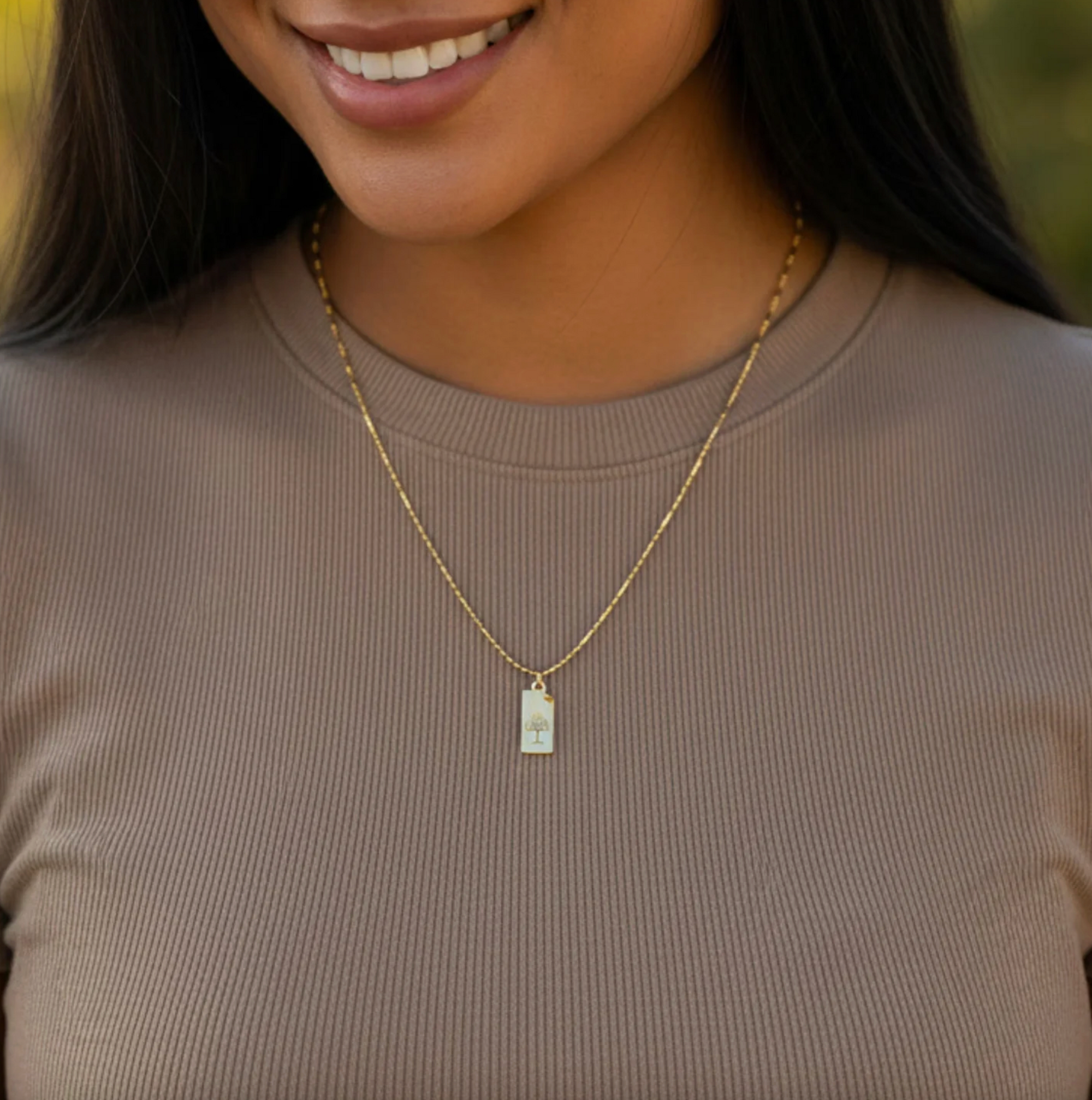 Woman wearing a gold necklace with a rectangular pendant against a neutral background