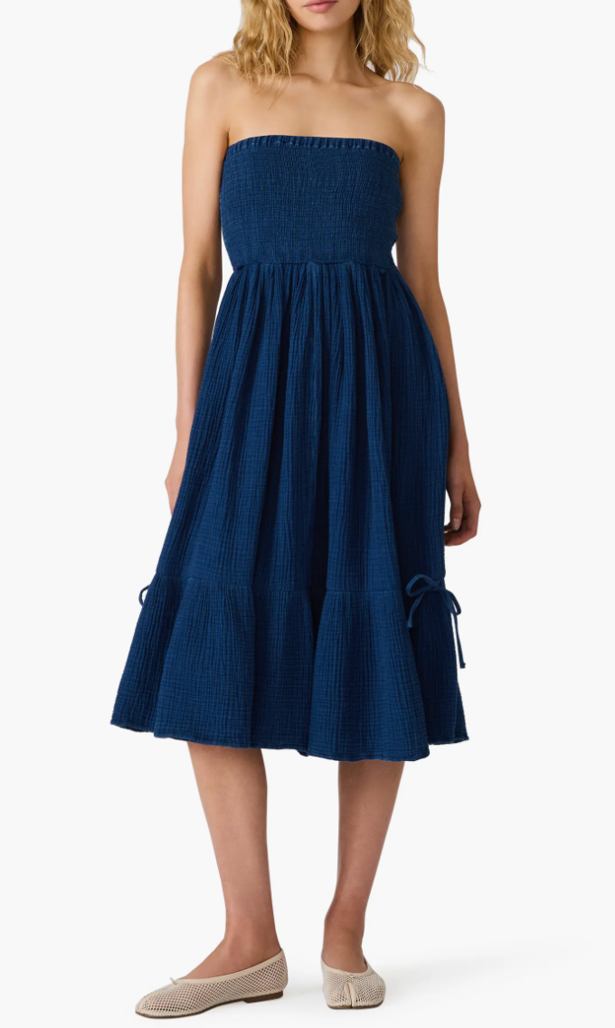 A woman modeling a strapless midi dress in dark blue with a textured, crinkled fabric. The dress features a smocked bodice and a flared skirt with side tie details, paired with beige flat shoes against a plain white background.