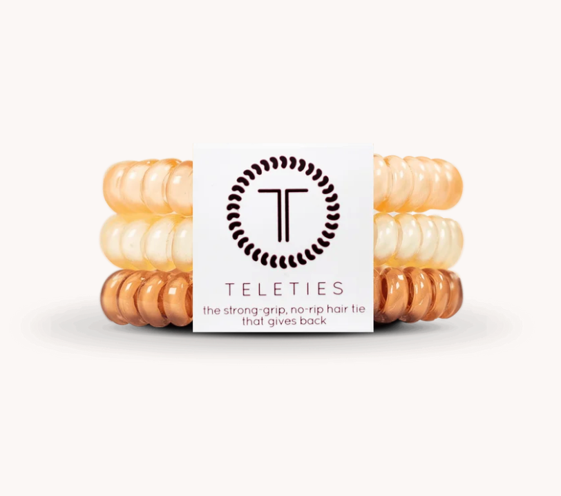 Three translucent spiral hair ties in peach, cream, and copper tones are stacked behind a white Teleties brand card on a clean white background.