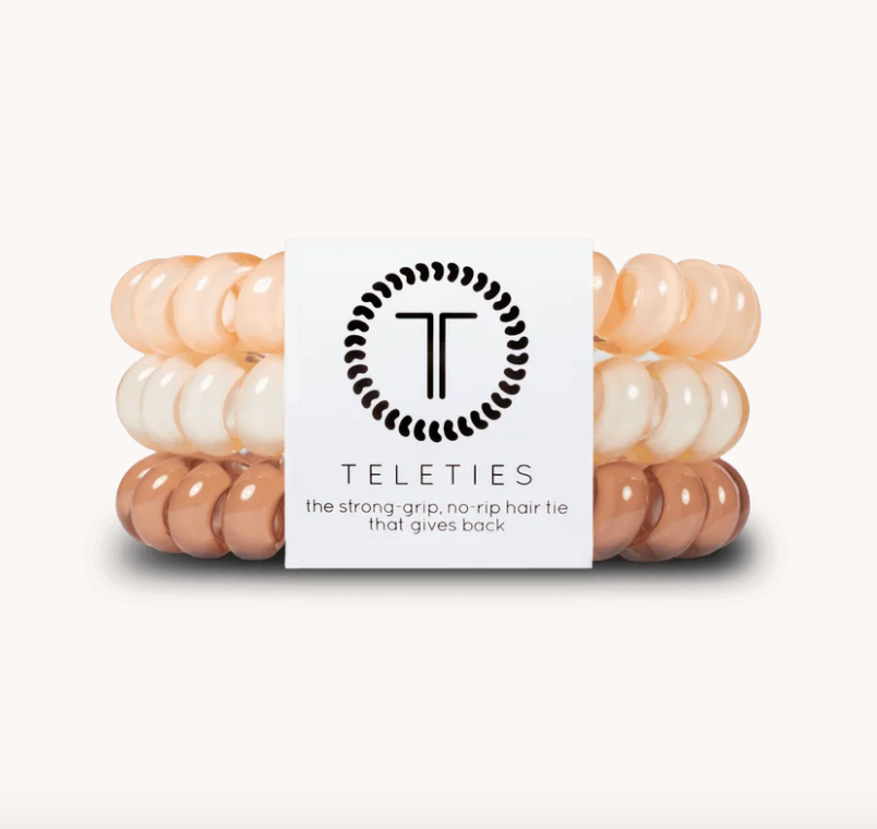 Three stacked Teleties hair ties in peach, cream, and copper tones, wrapped with a white paper band featuring the black Teleties logo and text.