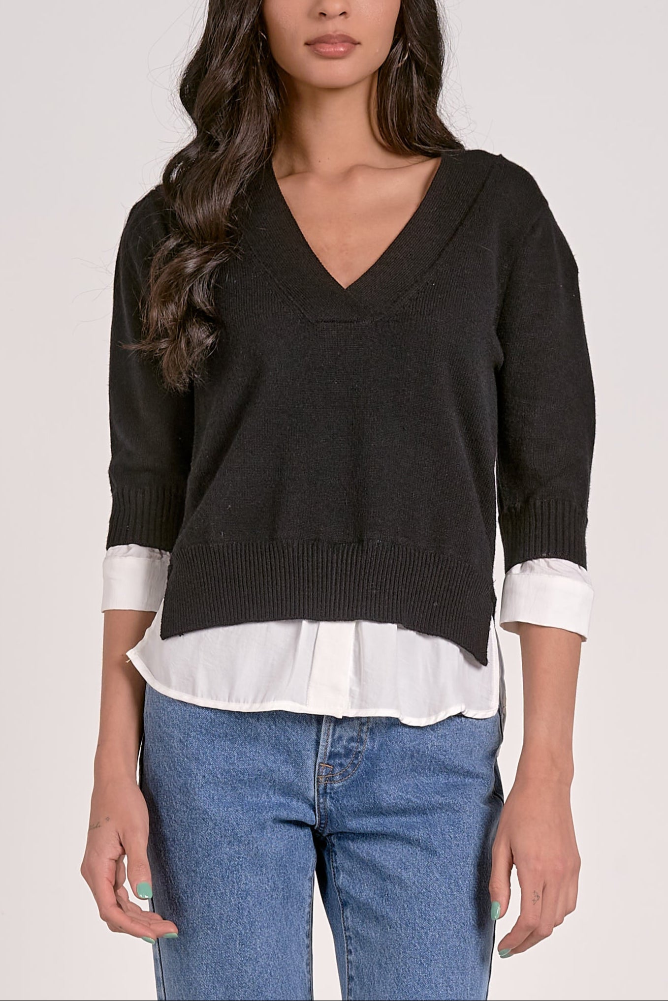 Woman wearing a black sweater over a white shirt with blue jeans on a plain background