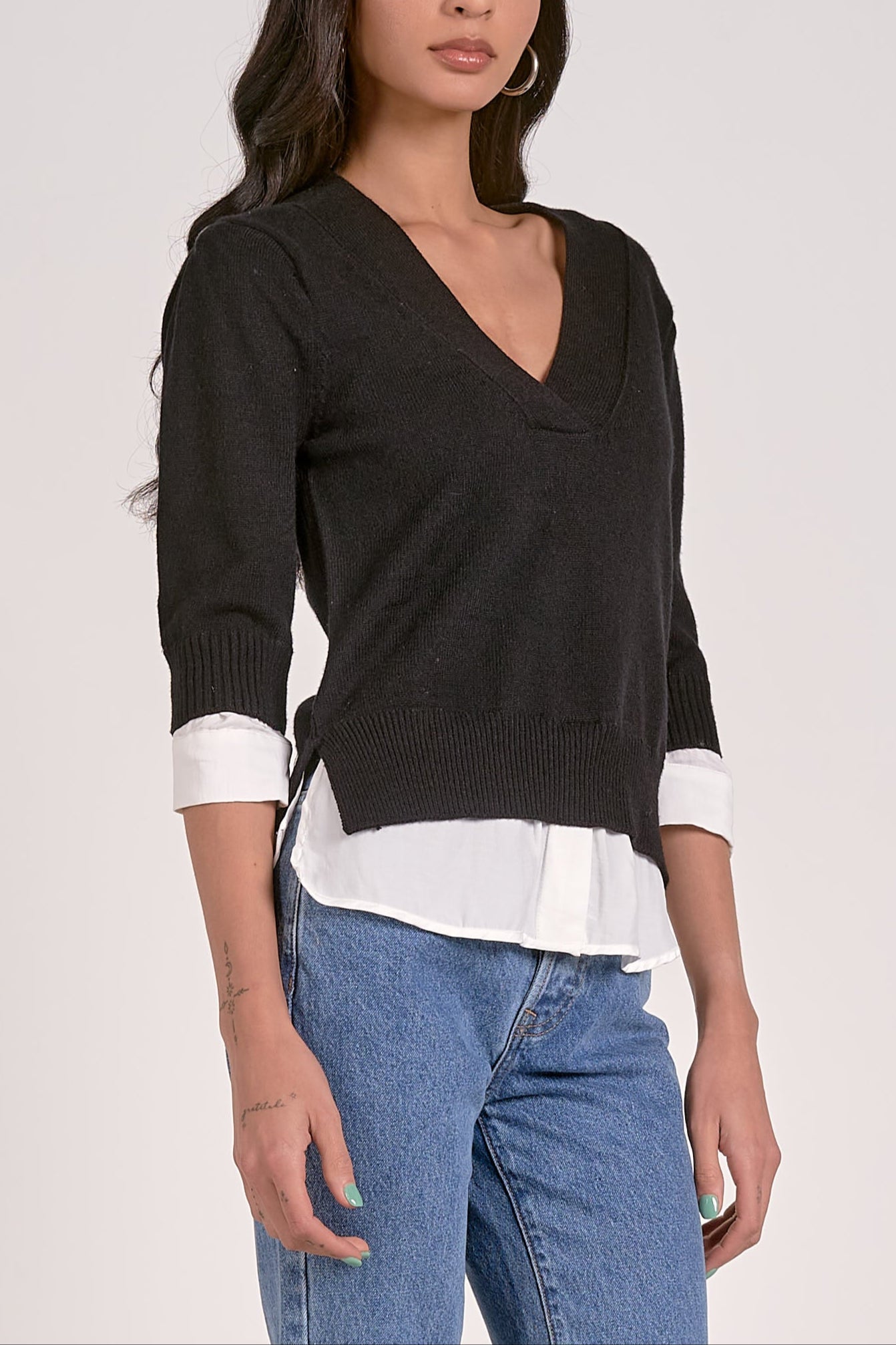 Woman wearing a black sweater over a white shirt with blue jeans on a light gray background